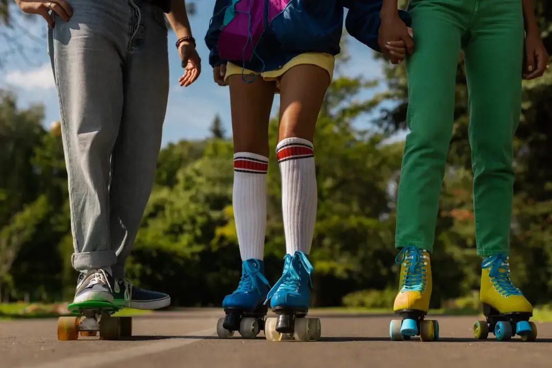 Drei Menschen nebeneinander auf einer Parkstraße: links auf einem Skateboard, in der Mitte und rechts auf Rollschuhen in Blau und Gelb